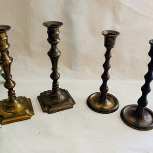 May include: Four antique brass and bronze candlesticks of varying designs. The candlesticks have ornate details and range in color from gold to dark bronze. The bases are square, round, and rectangular.