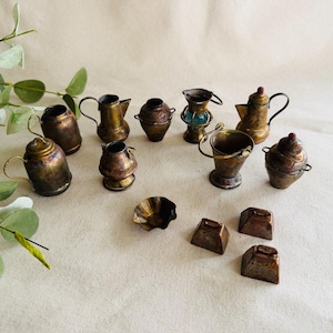 May include: A collection of antique miniature brass and copper kitchenware, including teapots, pitchers, and a small bucket. The pieces have a tarnished, aged appearance, with intricate details and handles. The arrangement is on a neutral-toned surface.