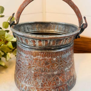 May include: An antique copper bucket with a hammered finish and intricate floral designs. The bucket has a copper handle with a hinged latch.