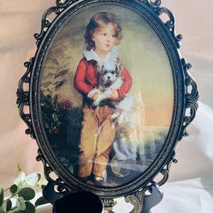 May include: An oval-shaped antique picture frame with a portrait of a child holding a dog. The frame is ornate, with a dark bronze finish. The child is wearing a red jacket and brown pants. A white mug with a black interior sits in front of the frame.