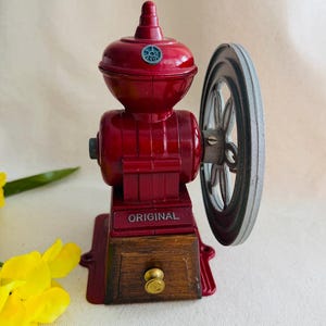 May include: A vintage red coffee grinder with a hand crank and a wooden drawer. The word "ORIGINAL" is printed on the front. The grinder has a wheel on the side and a small blue emblem on top.