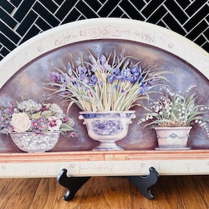 May include: A semi-circular decorative wall art piece featuring a floral still life. The artwork depicts three potted plants: irises, roses, and lily of the valley. The piece is supported by a black easel on a wooden surface.