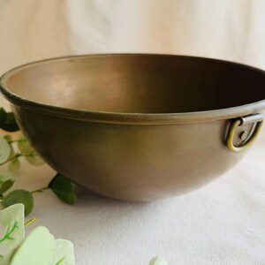 May include: A copper-colored bowl with a rounded shape and a small handle on the side. The bowl has a smooth surface and a slightly darker rim. The background is a soft, neutral color.