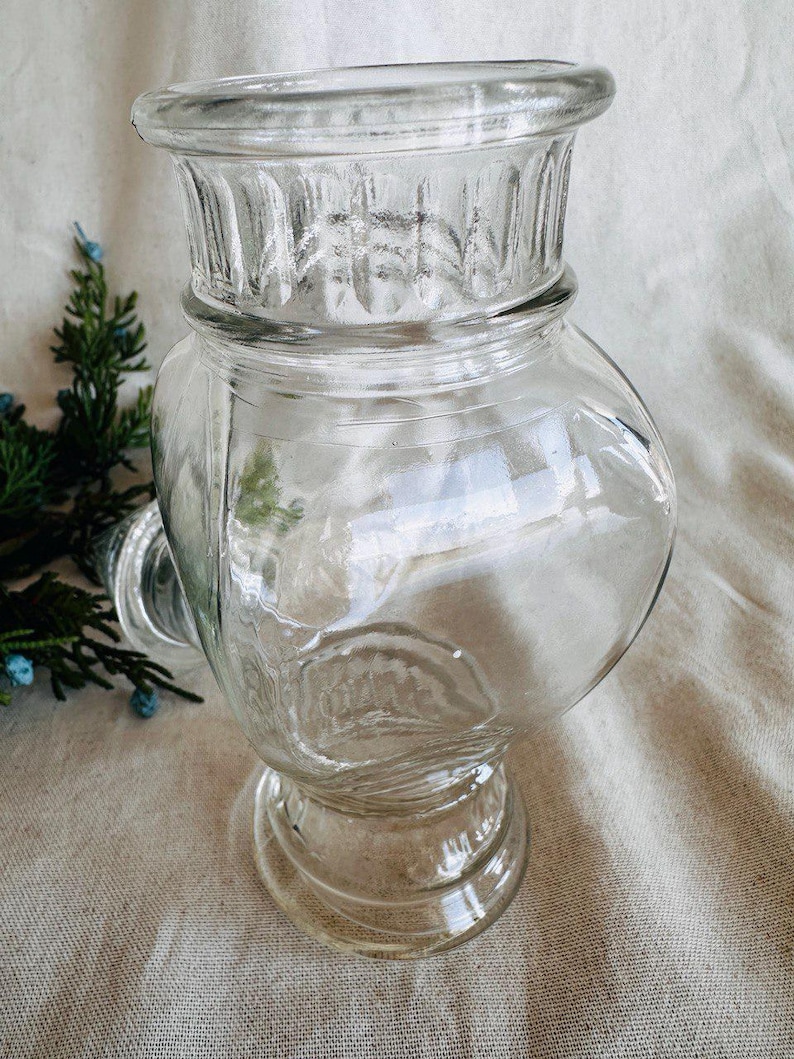 May include: A clear glass jar with a wide mouth and a textured design. The jar has a round base and a short neck.