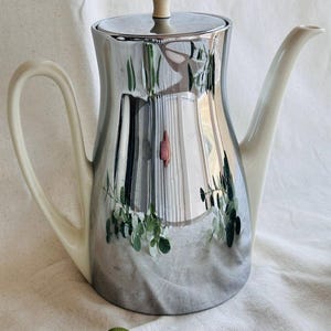 May include: A silver-colored teapot with a reflective surface, featuring a light-colored handle, spout, and lid. The teapot reflects a scene with green foliage and a pink flower. The lid has a small, light-colored knob.