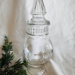 May include: A clear glass jar with a tall, pointed lid. The jar has a round body and a wide base. The lid has a decorative design of ridges and grooves.