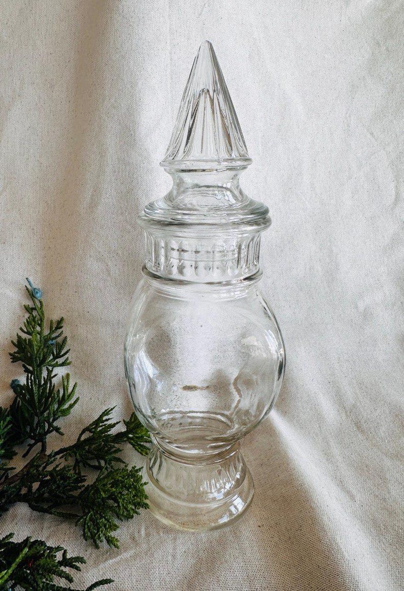 May include: A clear glass jar with a tall, pointed lid. The jar has a round body and a wide base. The lid has a decorative design of ridges and grooves.