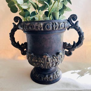 May include: A bronze-colored decorative urn with ornate handles and detailing. The urn is filled with green leafy stems. The urn has a pedestal base and is likely made of metal. The handles are curved and feature a leaf design.