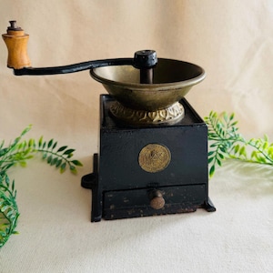 May include: Antique black coffee grinder with a brass bowl and a wooden handle. The grinder has a drawer and a decorative emblem on the front. The handle is attached to a black arm. The grinder is a vintage kitchen tool.