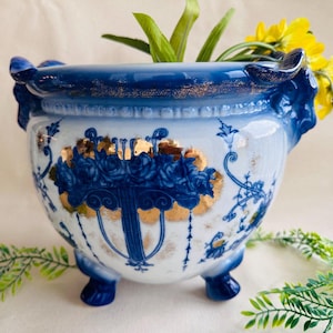 May include: A white and blue ceramic planter with a floral design and gold accents. The planter has three feet and decorative handles. Yellow flowers and green foliage are arranged inside the planter.
