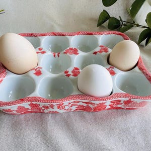 May include: A white ceramic egg holder with a red floral pattern holds three eggs. The egg tray has a scalloped edge and is designed to hold a dozen eggs. The eggs are different colors, with one white and two beige eggs.