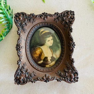 May include: Ornate brown oval picture frame with a portrait of a woman holding a dog. The frame has detailed floral and scrollwork designs. The portrait features a woman in a yellow and brown dress, with a dog in her arms.