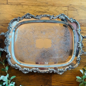 May include: Ornate silver-plated serving tray with a rectangular shape and decorative handles. The tray features an embossed floral pattern on the interior and a detailed, scrolling border. The tray is set against a wooden background.