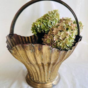 May include: A vintage brass basket with a handle, featuring a fluted design and a round base. The basket is filled with dried green and pink hydrangeas, creating a rustic and elegant display. The basket's aged patina adds to its antique charm.