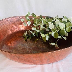 May include: A large, antique copper pan with two loop handles. The pan has a hammered texture and a rich, reddish-brown patina. Green and white eucalyptus branches rest inside the pan, adding a touch of natural decor.