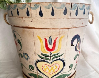 May include: A white wooden bucket with painted floral designs in red, yellow, blue, and green. The bucket has a heart shape with a cross-hatch pattern in the center. The top of the bucket has a blue and white painted border with black teardrop shapes.