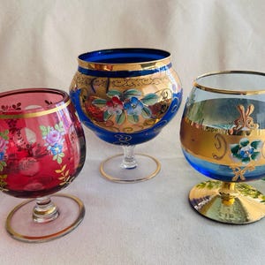 May include: Three ornate, hand-painted brandy snifters. The glasses feature gold rims and bases, with floral designs in red, blue, and gold. The glass on the left is red, the middle is blue, and the right is blue and gold.