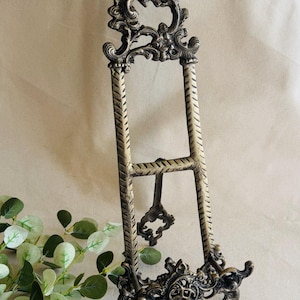 May include: Ornate, antique-style metal display easel in a bronze finish. The easel features decorative scrollwork at the top and base, with textured vertical supports. A small, decorative support extends from the back.