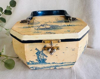 May include: Vintage hexagonal box with a blue and white Dutch windmill scene. The box has a black handle and a brass clasp. The box is painted in a cream color with blue accents. The box is likely made of wood.