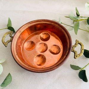 May include: A copper escargot dish with five circular indentations. The dish has two ornate brass handles and a polished finish. The escargot dish is a kitchenware item.