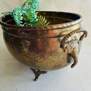 May include: A vintage brass planter with an aged patina. The bowl-shaped planter features an elephant head handle and three decorative feet. Green faux foliage is inside the planter. A decorative piece for home decor.