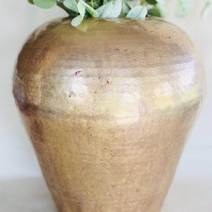 May include: A large, rounded brass vase with a textured, hammered surface finish. The metal has a warm, golden-brown patina with subtle variations in tone. A cluster of green eucalyptus stems with rounded leaves is placed inside the vase opening.