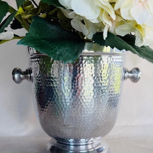 May include: A silver-toned, hammered metal ice bucket featuring a flared rim, a pedestal base, and two side handles. The bucket is filled with a bouquet of cream-colored hydrangea flowers with green leaves extending over the top edge.