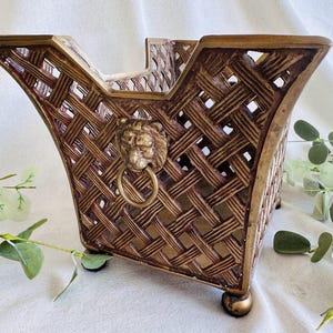 May include: A decorative, woven-style basket with a gold and brown finish. The basket features a lion's head with a ring handle on the front. The basket has a square base and is elevated by small, round feet. The basket is empty.