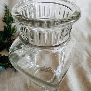 May include: A clear glass vase with a square base and a fluted top. The vase is made of thick glass and has a classic design.