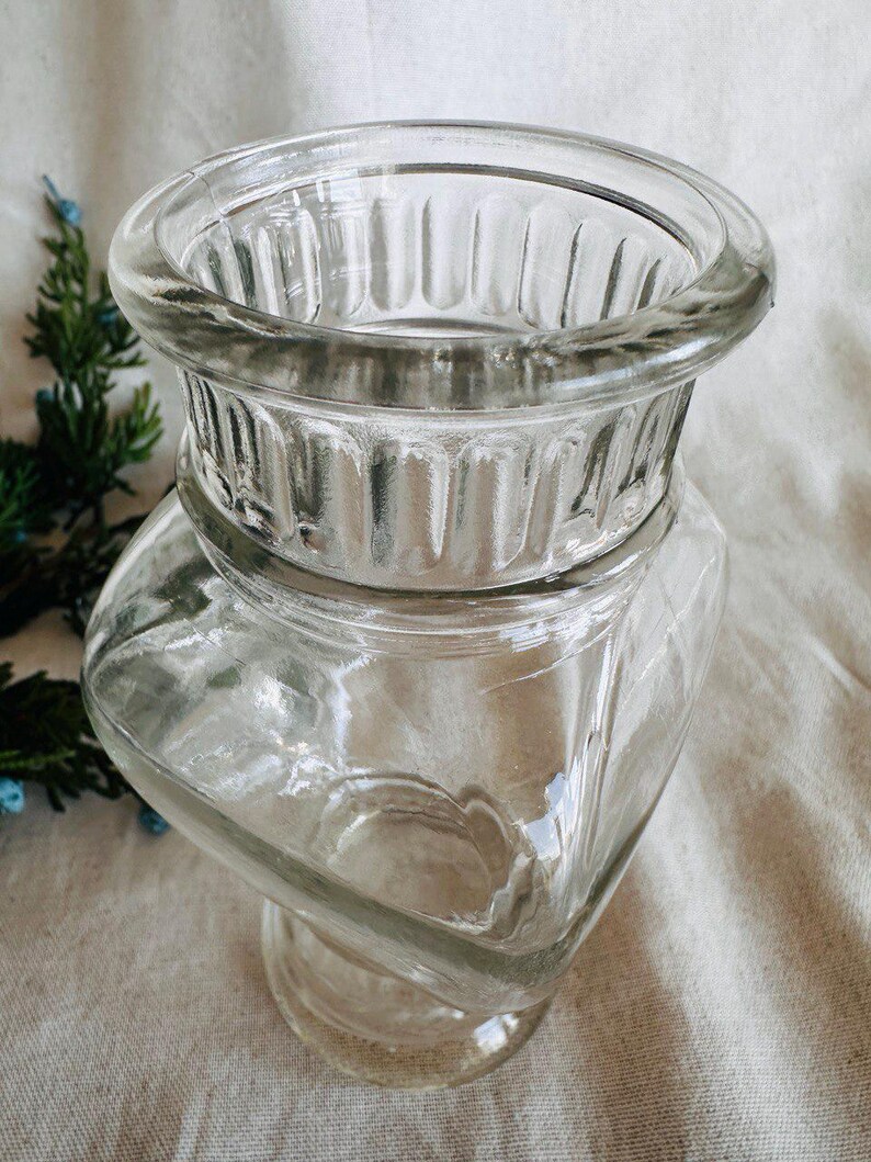 May include: A clear glass vase with a square base and a fluted top. The vase is made of thick glass and has a classic design.