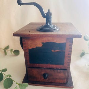 May include: An antique wooden coffee grinder with a dark finish. The grinder features a metal crank handle and a small drawer. The wooden body has a dark stain and a black painted section. The grinder sits on a light-colored surface.