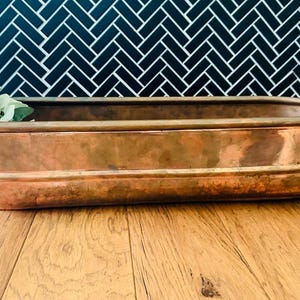 May include: An elongated, oval-shaped copper planter with a smooth, metallic finish and a slightly weathered patina. The vessel features a reinforced rim and a horizontal band detail along the side, containing a small green leafy plant.