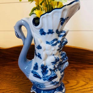 May include: A white and blue ceramic pitcher vase with a swan-shaped handle and a detailed, embossed design. The vase is filled with yellow artificial flowers. The vase is on a wooden surface.