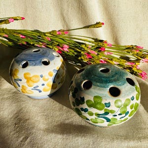 May include: Two ceramic flower vases with floral patterns. One vase is blue and yellow with a white background. The other vase is green and white with a white background.