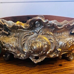 May include: A gold-toned metal planter with intricate floral designs and scrollwork. The planter has a rectangular shape with a curved top and four legs.