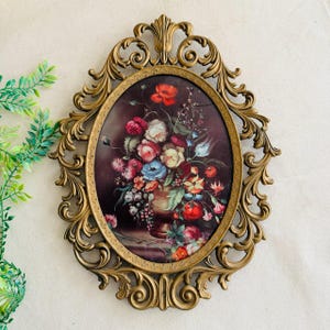 May include: An oval-shaped, antique gold-colored frame with an ornate design. The frame surrounds a floral still life painting featuring a bouquet of colorful flowers in a vase. The flowers include red, pink, white, and blue blooms.