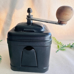 May include: A black manual coffee grinder with a wooden handle and a metal crank. The grinder has a square base and a small opening on the front. The wooden handle is round and light brown.