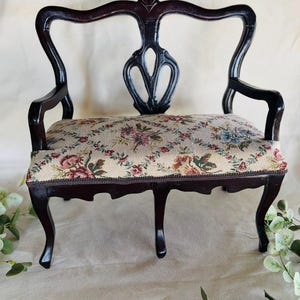 May include: A miniature dark brown wooden settee featuring a curved backrest with a central cutout design and cabriole legs. The seat is upholstered in a beige fabric with a repeating floral pattern in shades of pink, red, blue, and green.