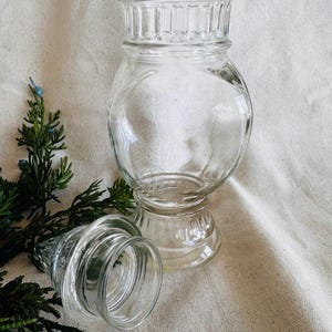 May include: A clear glass jar with a wide mouth and a decorative, fluted lid. The jar has a round body and a pedestal base. The jar is empty and appears to be vintage.