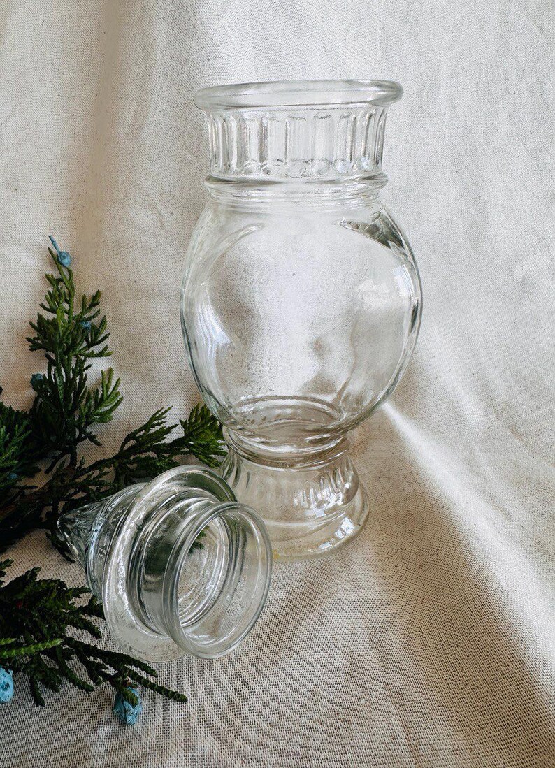 May include: A clear glass jar with a wide mouth and a decorative, fluted lid. The jar has a round body and a pedestal base. The jar is empty and appears to be vintage.