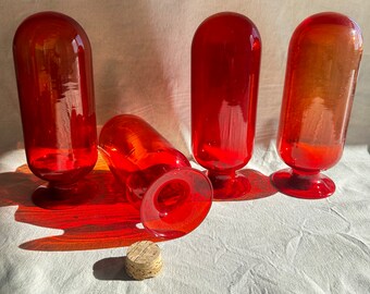 May include: Four red glass bottles with a cork stopper. The bottles have a rounded top and a flared base.