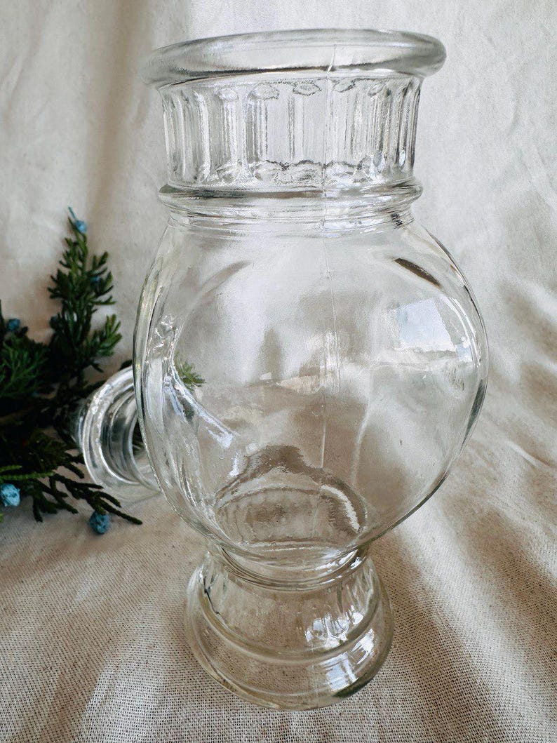 May include: A clear glass jar with a wide mouth and a decorative base. The jar has a fluted rim and a rounded body.