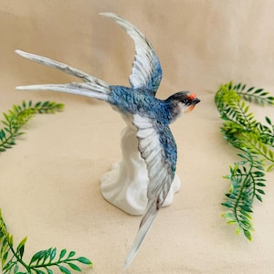 May include: A porcelain figurine of a swallow in flight. The bird has blue and white wings, a gray head with an orange spot, and a long, forked tail. It is perched on a white cloud-shaped base. The figurine is a decorative item.