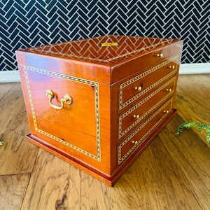 May include: A wooden jewelry box with a glossy finish. The box is a warm reddish-brown color with gold accents. It has a handle and several drawers. The box sits on a wooden surface.