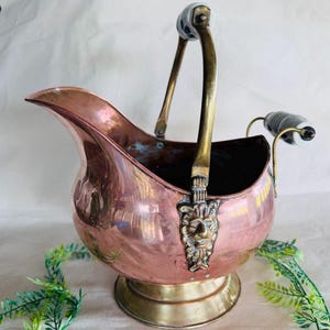 May include: A copper coal scuttle with brass accents and a lion's head emblem. The handle has white and blue ceramic details. The scuttle has a rounded body and a spout, and sits on a brass base. A decorative piece.