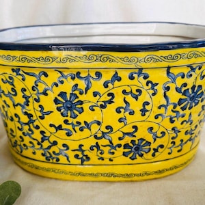 May include: A yellow ceramic planter with blue floral designs and two handles. The planter is oval shaped and has a white interior.