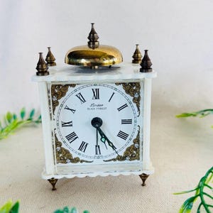 May include: A square white table clock with a brass bell on top and four brass finials at the corners. The clock face features black Roman numerals, black hands, and the text Linden Black Forest. The frame has gold-toned decorative corner accents.
