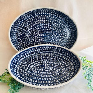 May include: Two oval-shaped ceramic serving dishes. The dishes have a dark blue background with a white dotted pattern. The rims are white. The dishes are nested on a light-colored surface.