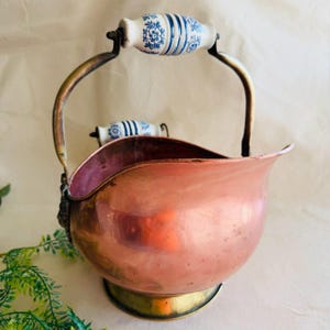 May include: A copper coal scuttle with a brass base and handle supports. The handle features a white ceramic grip with blue floral and striped designs. The scuttle has a curved shape and is likely vintage.
