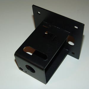 May include: A black metal bracket with a rectangular base and a box-like structure attached at an angle. The bracket has several holes and cutouts, and is likely used for mounting or support.